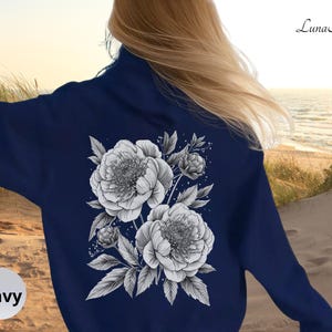 K&ouml;nnte beinhalten: Marineblaues Sweatshirt mit einem schwarz-wei&szlig;en Blumendesign auf dem R&uuml;cken, das Pfingstrosen und Bl&auml;tter zeigt. Das Wort "Navy" befindet sich in einem wei&szlig;en Kreis unten links. Der Markenname "Luna Alla" steht oben rechts.