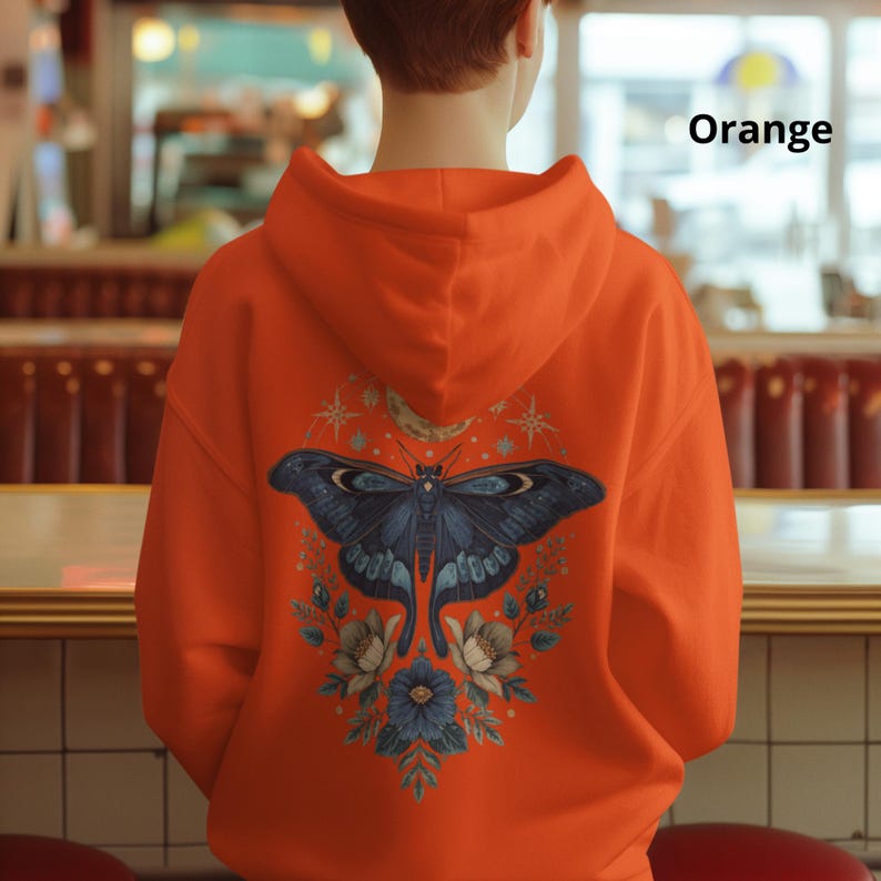 May include: An orange hoodie featuring a large graphic on the back. The design displays a dark blue moth centered above floral elements, a crescent moon, and star shapes. The word Orange is printed in black text in the upper right corner.
