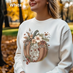 Puede incluir: Una sudadera blanca de cuello redondo con un gráfico central de una gran calabaza naranja adornada con flores, hojas y elementos botánicos en tonos tierra apagados. La palabra White está impresa en la esquina superior izquierda.