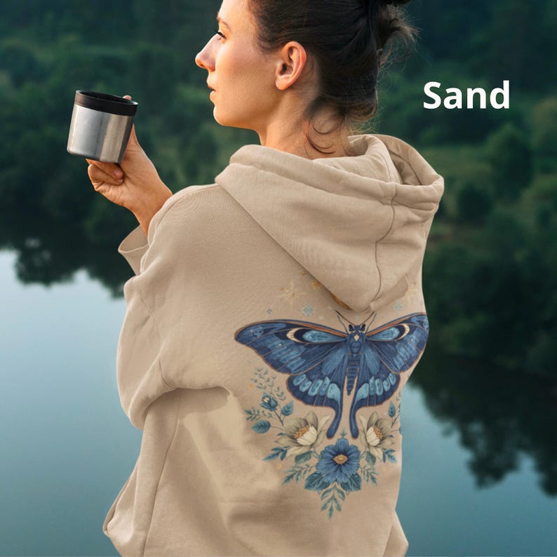 May include: A sand-colored hooded sweatshirt featuring a large graphic on the back of a blue moth surrounded by blue and white flowers and small stars. The word Sand is printed in white text in the upper right corner of the image.