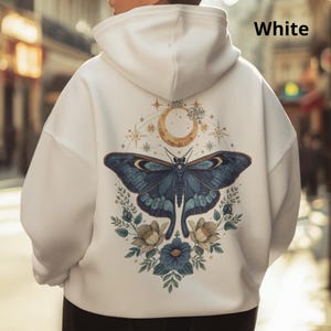 May include: A white hooded sweatshirt featuring a large graphic on the back. The design displays a dark blue moth centered below a golden crescent moon and stars, surrounded by blue and beige floral elements. The word White is printed in the corner.