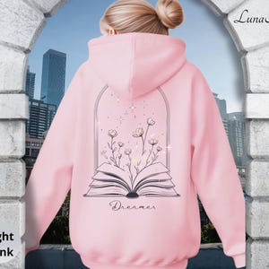 May include: A light pink hooded sweatshirt featuring a black line art graphic on the back. The design depicts an open book with wildflowers growing from the pages, framed by an archway with small stars. The word Dreamer is printed below the book.
