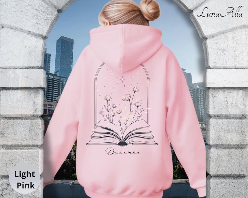 May include: A light pink hooded sweatshirt featuring a black line art graphic on the back. The design depicts an open book with wildflowers growing from the pages, framed by an archway with small stars. The word Dreamer is printed below the book.