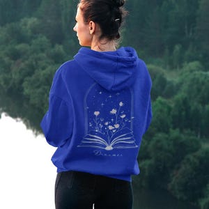 May include: A royal blue hooded sweatshirt featuring a white line art graphic on the back. The design depicts an open book with flowers growing from the pages inside an arched frame, accompanied by stars and the word Dreamer written in script.