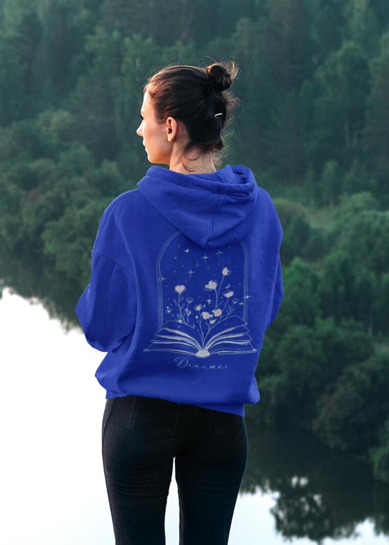 May include: A royal blue hooded sweatshirt featuring a white line art graphic on the back. The design depicts an open book with flowers growing from the pages inside an arched frame, accompanied by stars and the word Dreamer written in script.