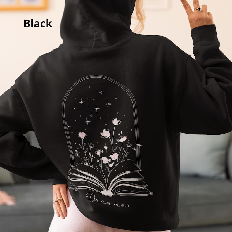 May include: A black hooded sweatshirt featuring a white line art graphic on the back. The design depicts an open book with flowers growing from the pages inside an arched frame, accented by stars. The word Dreamer is printed below the book.