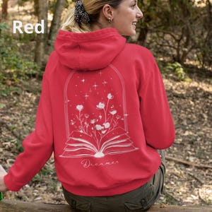 May include: A red hooded sweatshirt featuring a white line art graphic on the back. The design depicts an open book with flowers growing from the pages, enclosed in an arched frame with stars. The word Dreamer is printed below the book.