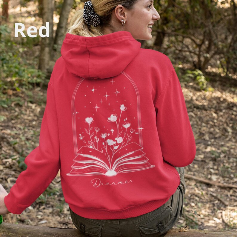 May include: A red hooded sweatshirt featuring a white line art graphic on the back. The design depicts an open book with flowers growing from the pages, enclosed in an arched frame with stars. The word Dreamer is printed below the book.