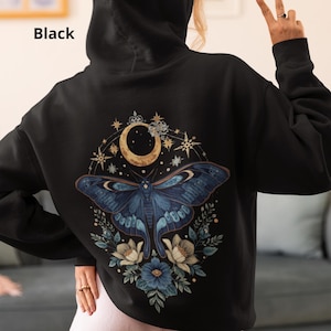 May include: A black hooded sweatshirt featuring a large graphic on the back. The design displays a detailed blue moth centered beneath a golden crescent moon, surrounded by stars and blue and white floral elements. The word Black is printed.