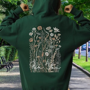 May include: A dark green hoodie with a floral design on the back. The design features various flowers and foliage in shades of white, beige, and brown, with the words "For You" in a cursive font. The hoodie has a hood and long sleeves.