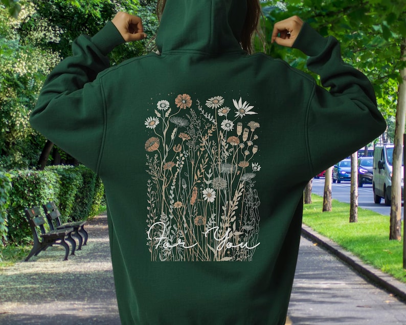 May include: A dark green hoodie with a floral design on the back. The design features various flowers and foliage in shades of white, beige, and brown, with the words "For You" in a cursive font. The hoodie has a hood and long sleeves.