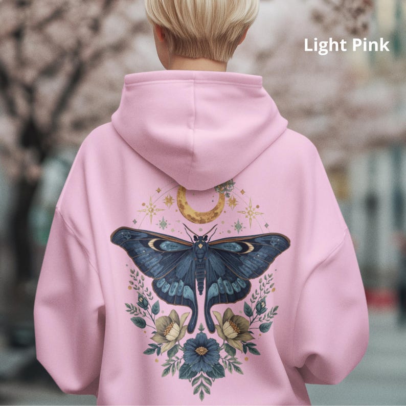 May include: A light pink hooded sweatshirt featuring a large graphic on the back. The design displays a dark blue moth centered below a golden crescent moon and stars, surrounded by blue and cream-colored flowers and green foliage.