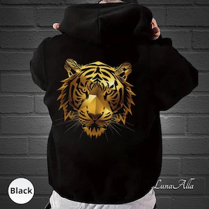 Può includere: Una felpa con cappuccio nera caratterizzata da una grande grafica centrale di una testa di tigre in uno stile geometrico color oro metallizzato. Il design della tigre presenta linee angolari nette e baffi dettagliati sul tessuto nero.