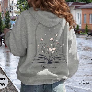 May include: A sport grey hooded sweatshirt featuring a graphic on the back of an open book with pink flowers growing out of the pages inside an arched frame. The word Dreamer is printed in a cursive script below the book illustration.