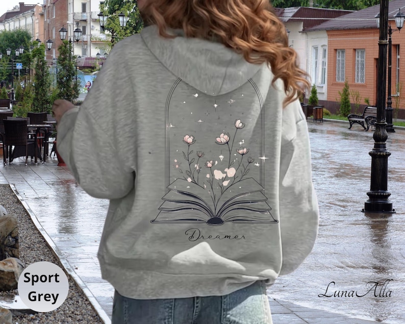 May include: A sport grey hooded sweatshirt featuring a graphic on the back of an open book with pink flowers growing out of the pages inside an arched frame. The word Dreamer is printed in a cursive script below the book illustration.