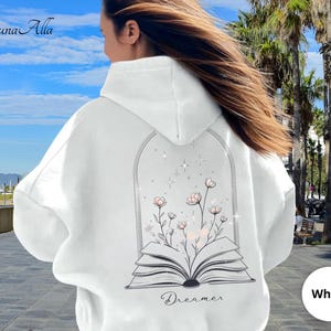 May include: A white hooded sweatshirt featuring a black line art graphic on the back. The design depicts an open book with flowers growing from the pages, enclosed within an arched frame with small star accents. The word Dreamer is printed below.