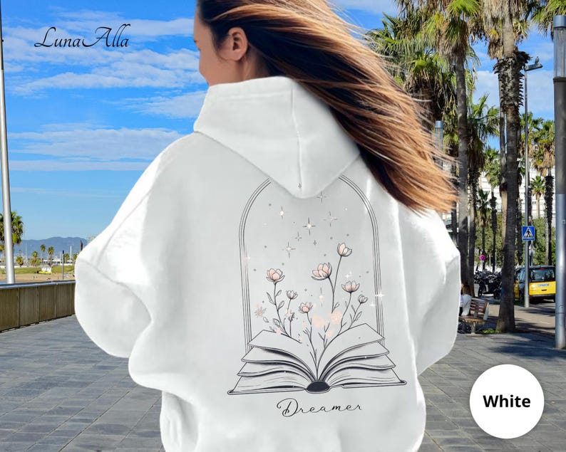 May include: A white hooded sweatshirt featuring a black line art graphic on the back. The design depicts an open book with flowers growing from the pages, enclosed within an arched frame with small star accents. The word Dreamer is printed below.