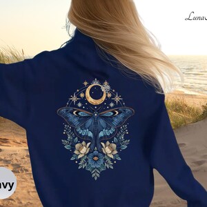 May include: A navy blue long-sleeve sweatshirt featuring a large back graphic of a moth centered beneath a crescent moon, surrounded by stars and floral elements. The text "Luna Alla" is printed in the upper right corner of the garment.