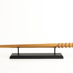May include: A light brown wooden wand with a carved handle and tapered tip, displayed on a black stand. The wand's handle features a series of rounded ridges. The stand is rectangular with two vertical supports.