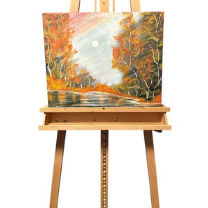 Könnte beinhalten: Ein Herbstlandschaftsgemälde auf Leinwand, ausgestellt auf einer Holzstaffelei. Das Kunstwerk zeigt Bäume mit orangefarbenem und gelbem Laub, die einen Fluss säumen, mit einem blassen Mond am Himmel. Die Staffelei ist aus hellem Holz.