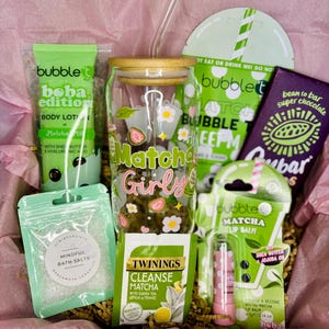 May include: A gift box filled with matcha-themed products. Includes a glass tumbler with a bamboo lid, lip balm, body lotion, bath salts, tea, and a chocolate bar. The tumbler has the words "Matcha Girly" and strawberry and flower designs.