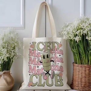 May include: Cream-colored tote bag with the words "ICED Matcha Club" in a retro font. The design includes a cartoon matcha drink with a straw, winking and giving a thumbs up. The bag has long handles.