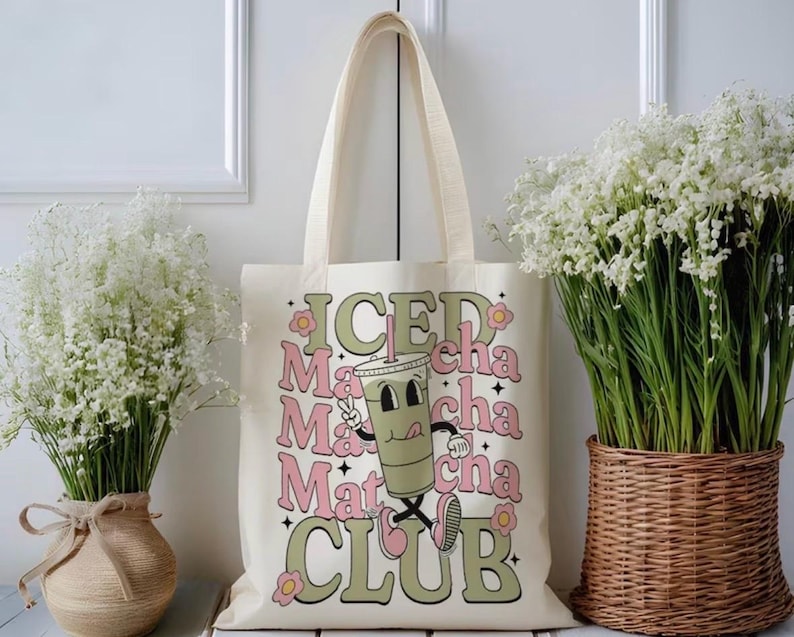 May include: Cream-colored tote bag with the words "ICED Matcha Club" in a retro font. The design includes a cartoon matcha drink with a straw, winking and giving a thumbs up. The bag has long handles.