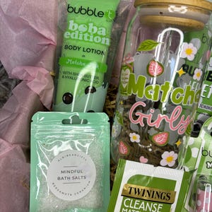 May include: A gift basket with self-care products. Includes a green tube of Bubble T Boba Edition Matcha Milk body lotion, a clear jar with a wooden lid, a bag of bath salts, and a Twinings Cleanse Matcha tea bag. The jar has a "Matcha Girly" design.