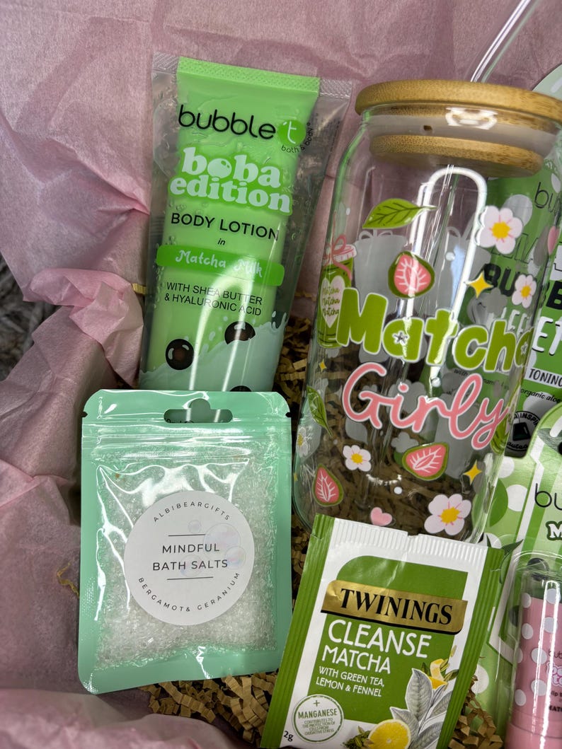 May include: A gift basket with self-care products. Includes a green tube of Bubble T Boba Edition Matcha Milk body lotion, a clear jar with a wooden lid, a bag of bath salts, and a Twinings Cleanse Matcha tea bag. The jar has a "Matcha Girly" design.