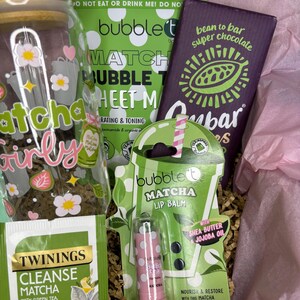 May include: A gift box filled with matcha-themed products. Includes a glass jar with floral decals, a matcha lip balm, a chocolate bar, a matcha sheet mask, and a tea bag. The box is lined with pink tissue paper and shredded paper.