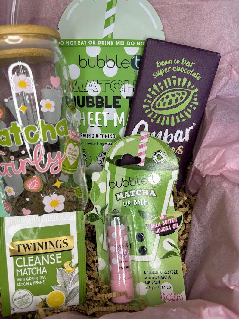 May include: A gift box filled with matcha-themed products. Includes a glass jar with floral decals, a matcha lip balm, a chocolate bar, a matcha sheet mask, and a tea bag. The box is lined with pink tissue paper and shredded paper.