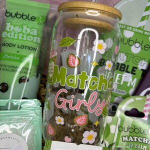 May include: A gift set featuring a glass jar with "Matcha Girly" text and floral designs, a body lotion tube, lip balm, and tea packets. The jar has a wooden lid and a straw. The set includes matcha-themed products.