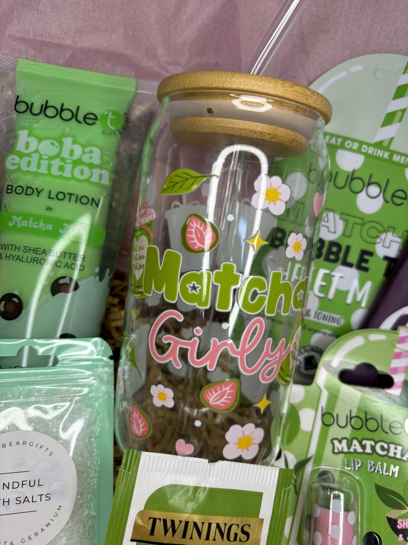 May include: A gift set featuring a glass jar with "Matcha Girly" text and floral designs, a body lotion tube, lip balm, and tea packets. The jar has a wooden lid and a straw. The set includes matcha-themed products.