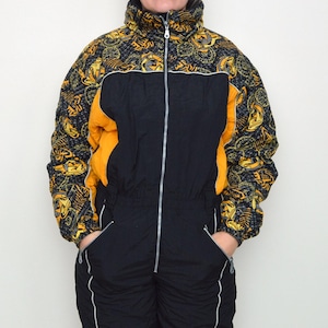 Könnte beinhalten: Vintage Ski-Overall in Schwarz und Gelb mit durchgehendem Reißverschluss. Die Ärmel haben ein gemustertes Design in Gelb und Schwarz mit orangefarbenen Akzenten an den Seiten. Der Anzug hat Taschen und weiße Zierleisten.