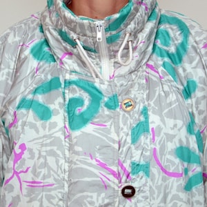 Puede incluir: Chaqueta de esquí vintage con un estampado abstracto gris, turquesa y morado. La chaqueta tiene un cuello alto con cremallera y cordón, y presenta dos botones. La chaqueta es de estilo retro.
