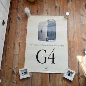Könnte beinhalten: Ein rechteckiges, cremefarbenes Poster mit einem Power Macintosh G4 Computer, dem Text "Introducing Power Macintosh G4 with Velocity Engine", einer großen "G4" Grafik und dem Apple-Logo mit dem Text "Think different" am unteren Rand.