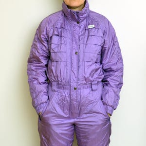 Puede incluir: Mono de esquí morado brillante vintage. El mono de una pieza tiene cuello alto, botones y bolsillos. El traje está acolchado y tiene cinturón en la cintura. El estilo general recuerda a la ropa de esquí de los años 80.