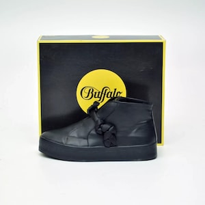 Puede incluir: Zapatilla alta negra con suela gruesa. El zapato presenta un detalle anudado en el lateral y está colocado frente a una caja negra con un círculo amarillo y la palabra "Buffalo".