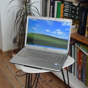 Könnte beinhalten: Ein silberner Dell-Laptop mit QWERTY-Tastatur und Touchpad. Auf dem Bildschirm ist das klassische Windows XP Bliss-Hintergrundbild zu sehen, das einen hellblauen Himmel mit weißen Wolken über einer grünen Hügellandschaft zeigt.