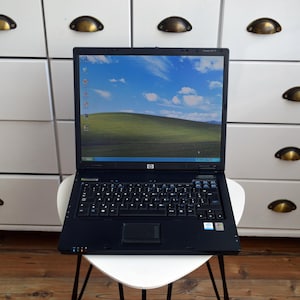 May include: A black HP laptop with a QWERTY keyboard and a trackpad. The screen displays the classic Windows XP Bliss wallpaper featuring a green hill under a blue sky with white clouds. The laptop is open and positioned on a white surface.
