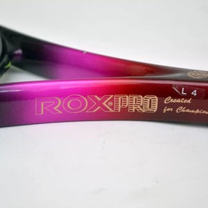 Könnte beinhalten: Nahaufnahme eines Tennisschlägers mit einem schwarzen und verlaufenden rosa und roten Rahmen. Der Text "ROXPRO" ist goldfarben auf den Rahmen gedruckt. Der Griff ist schwarz und die Saiten sind gelb.