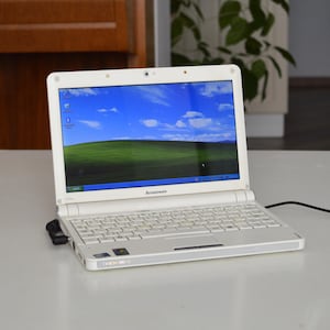 Könnte beinhalten: Ein weißer Lenovo-Laptop mit kompakter Tastatur und einem Bildschirm, der das klassische Windows XP Bliss-Hintergrundbild mit einem grünen Hügel und blauem Himmel zeigt. Das Gerät verfügt über ein schwarzes Stromkabel an der Seite.