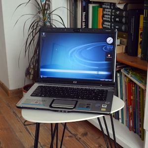 Pode incluir: Um computador portátil HP prateado e preto com um ecrã de ambiente de trabalho azul, um teclado preto e um touchpad prateado. O portátil está aberto, exibindo um relógio digital e ícones no ecrã, com uma câmara web no topo da moldura.