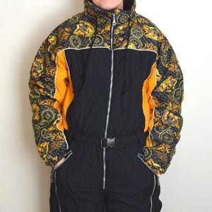 Puede incluir: Traje de esquí vintage negro y naranja con una sección superior estampada. El traje tiene una cremallera de largo completo, un cinturón y bolsillos. La sección estampada presenta diseños dorados y negros.