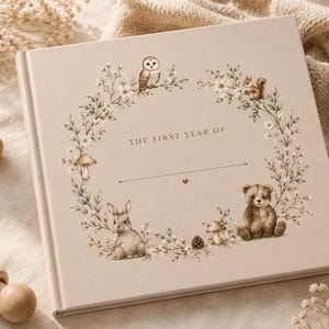 Puede incluir: Un libro de recuerdos para bebés de color beige claro con una ilustración de corona floral que incluye un búho, una ardilla, un conejo, un oso y setas. El texto de la portada dice THE FIRST YEAR OF seguido de una línea y un corazón.