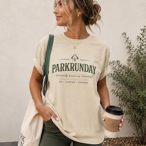May include: A beige short-sleeved t-shirt featuring a dark green graphic with a tree icon and the text PARKRUNDAY (previously known as Saturday) 5K • COFFEE • FRIENDS. The shirt is paired with dark green shorts and a beige canvas tote bag.