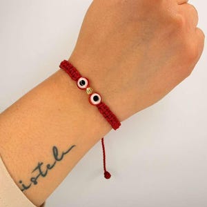 Op de afbeelding: Rode geweven armband met twee evil eye bedels met witte en zwarte details en een gouden kraal. De armband is vastgebonden met een rood touwtje. Het woord "isteb" is op de pols getatoeëerd.