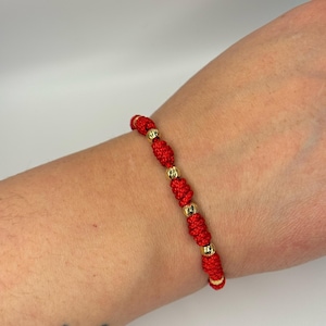 May include: Red knotted bracelet with gold-colored beads. The bracelet is made of a red cord with several gold-colored beads evenly spaced along the length. The bracelet is worn on a wrist.