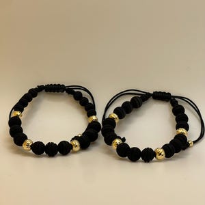 May include: Two black beaded bracelets with gold-colored accents. The bracelets feature a series of black beads interspersed with gold-colored, textured beads. The bracelets are on a white background.