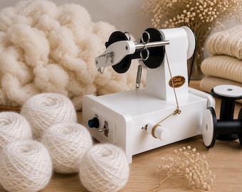 May include: A white electric yarn winder with black details and the text "Cozy Corner." The image also shows several balls of white yarn, a pile of fluffy white wool, and a stack of cream-coloured jumpers.